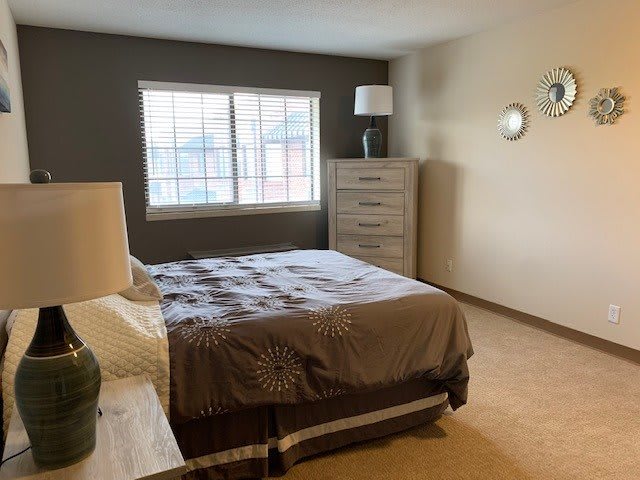 A cozy bedroom featuring a bed with a nightstand, a lamp, and a dresser by the window.