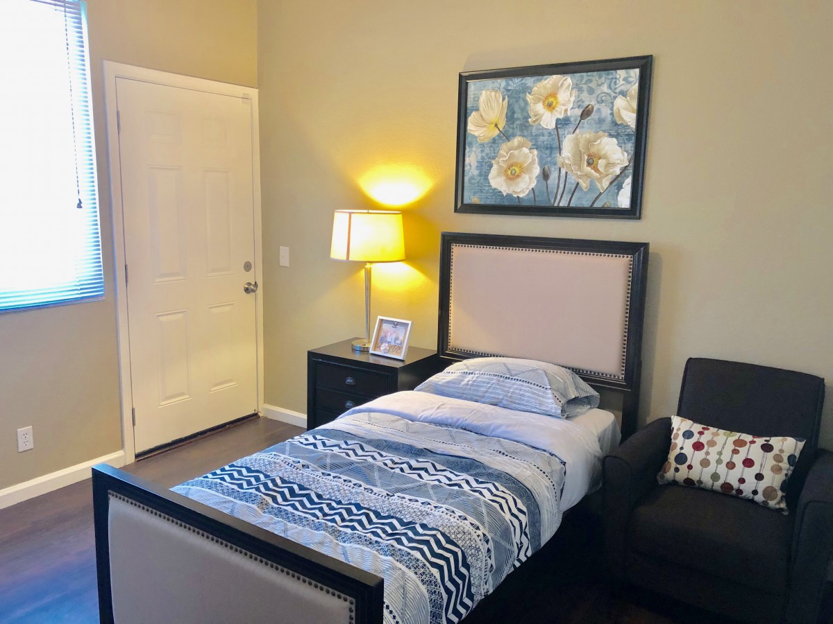 A cozy bedroom with a single bed, bedside table, lamp, and floral artwork on the wall.
