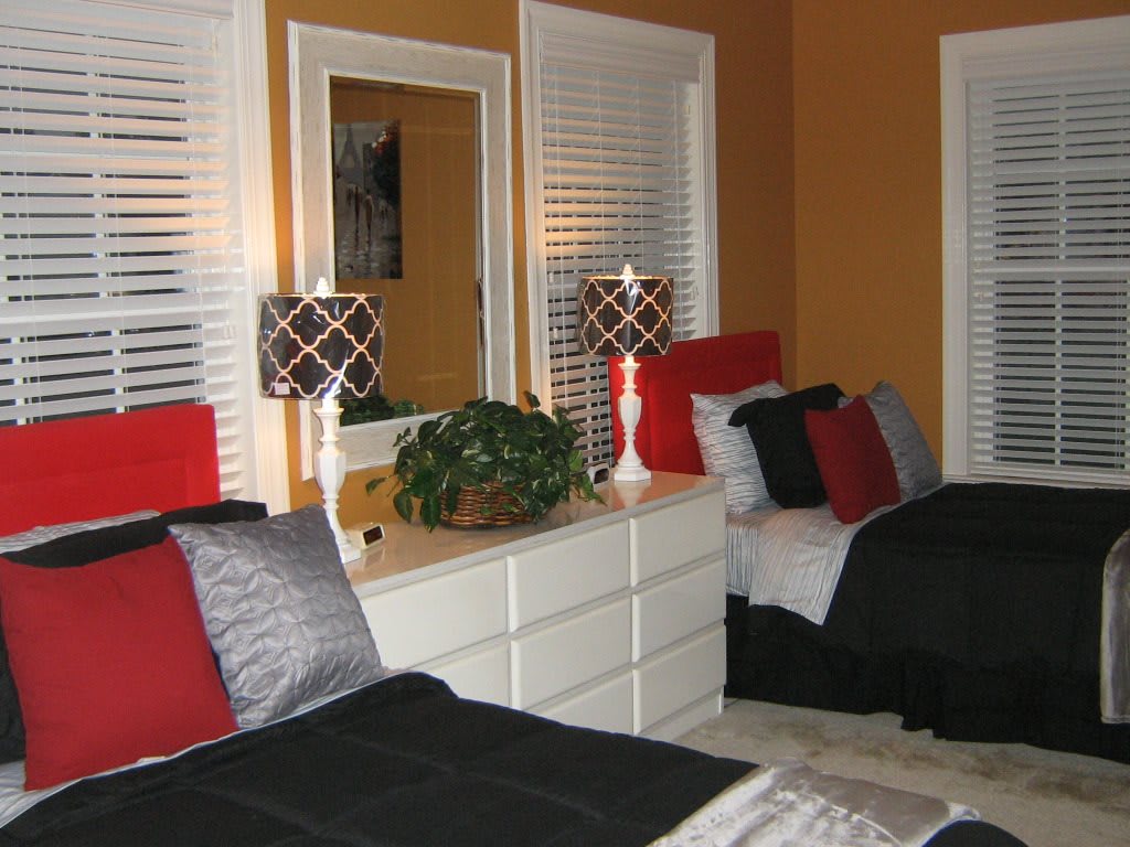 A modern bedroom with twin beds, featuring red and black pillows, matching lamps, and a central dresser with a mirror.