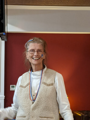 A smiling woman with glasses and a beaded necklace stands in a warmly colored interior.