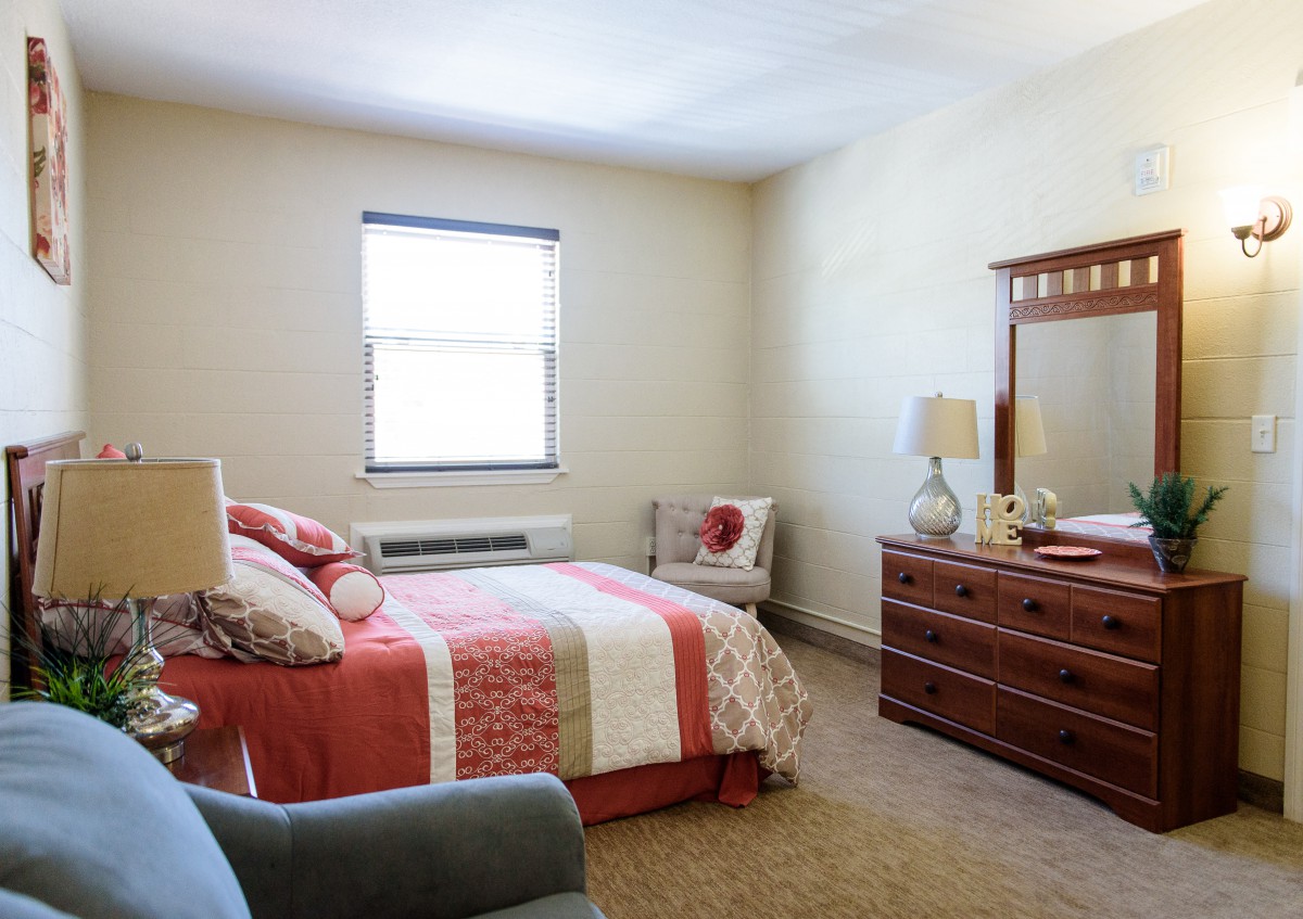 A cozy bedroom with a neatly made bed, a dresser with a mirror, and a chair by the window.