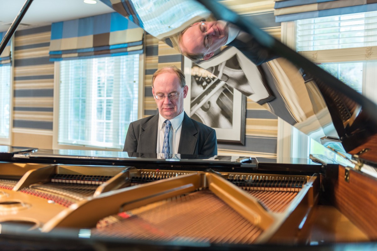 A man in a suit playing the piano indoors with his reflection visible.