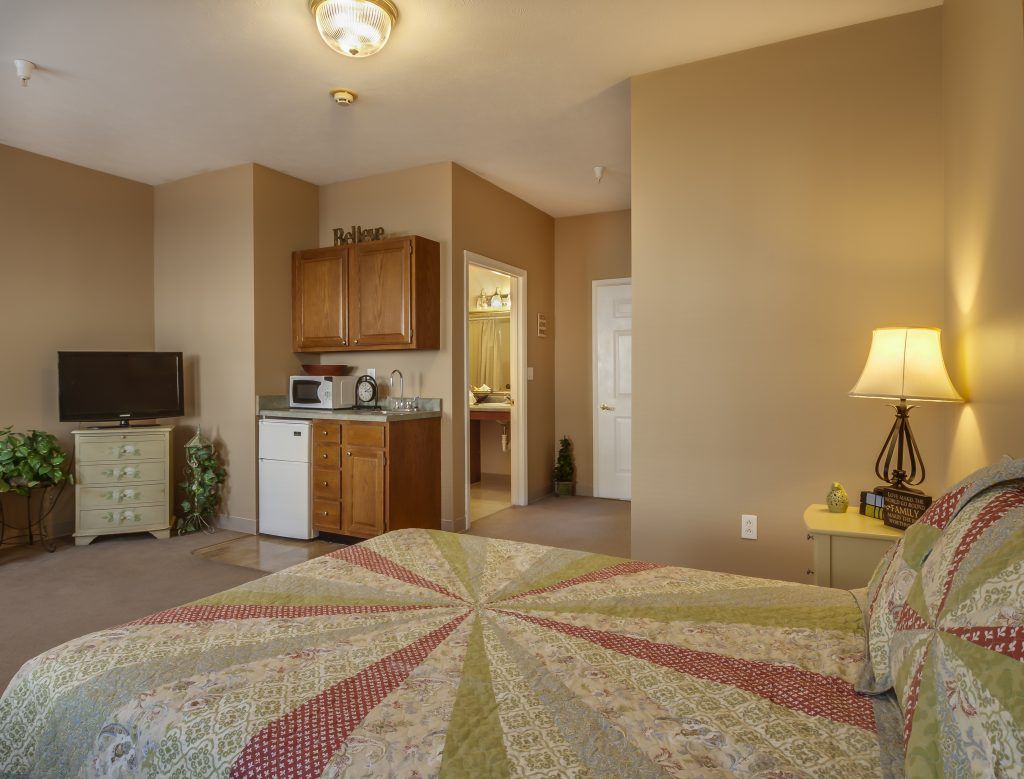 A cozy bedroom with a quilted bedspread, kitchenette, and TV.