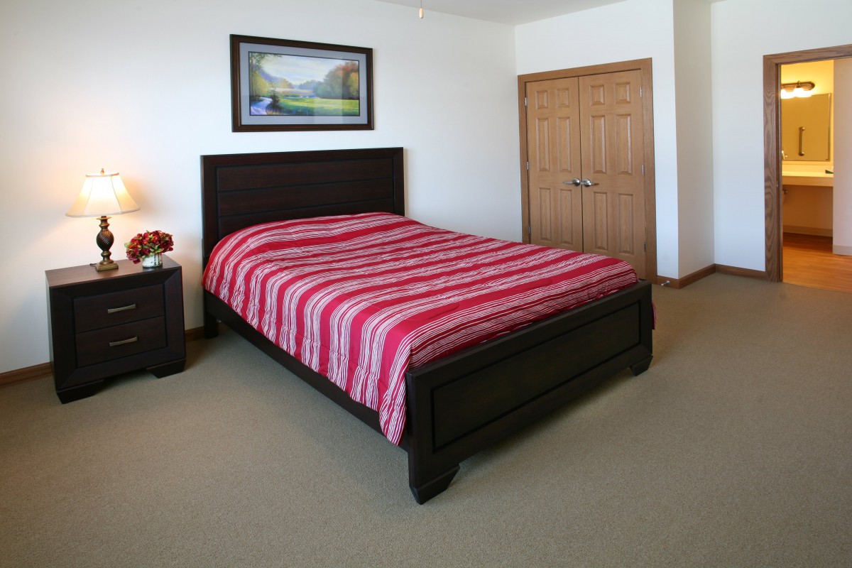 A neatly arranged bedroom featuring a bed with red striped bedding, a nightstand with a lamp, and a landscape painting on the wall.