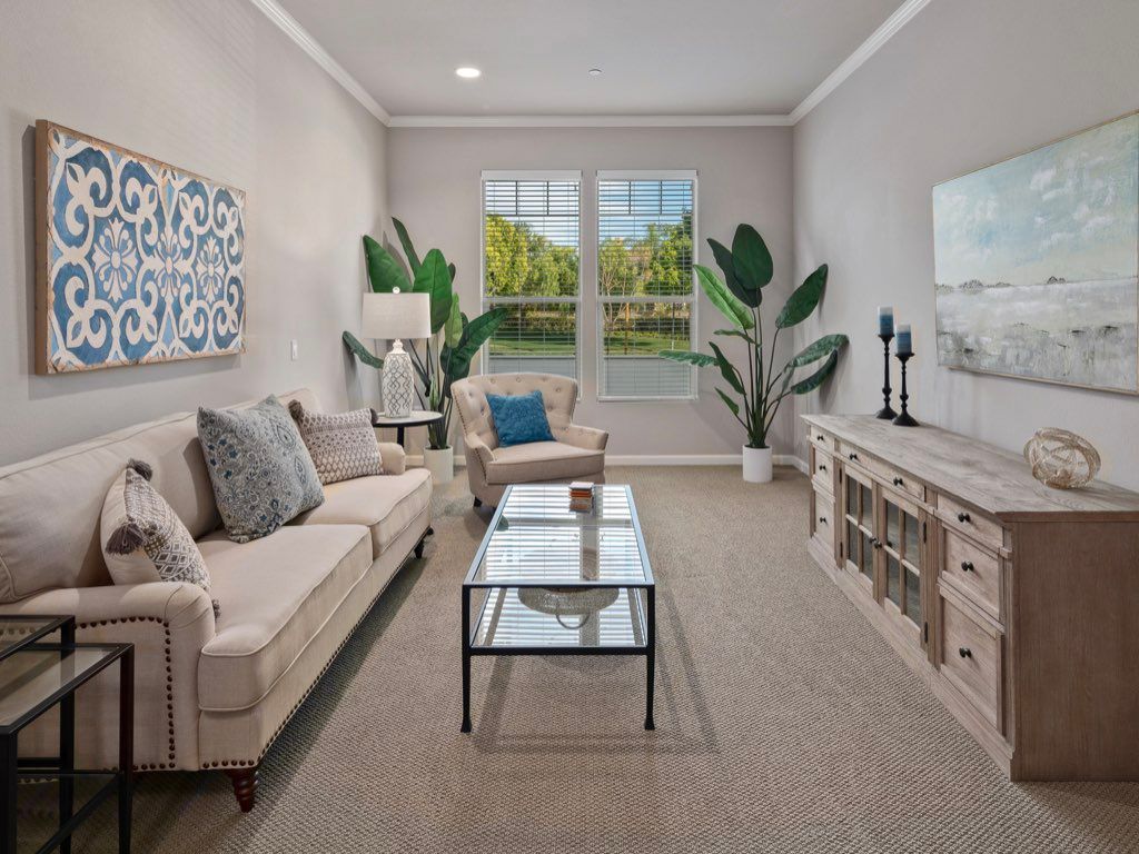 A modern living room with neutral tones, featuring a comfortable sofa, a glass coffee table, and large potted plants by the window.