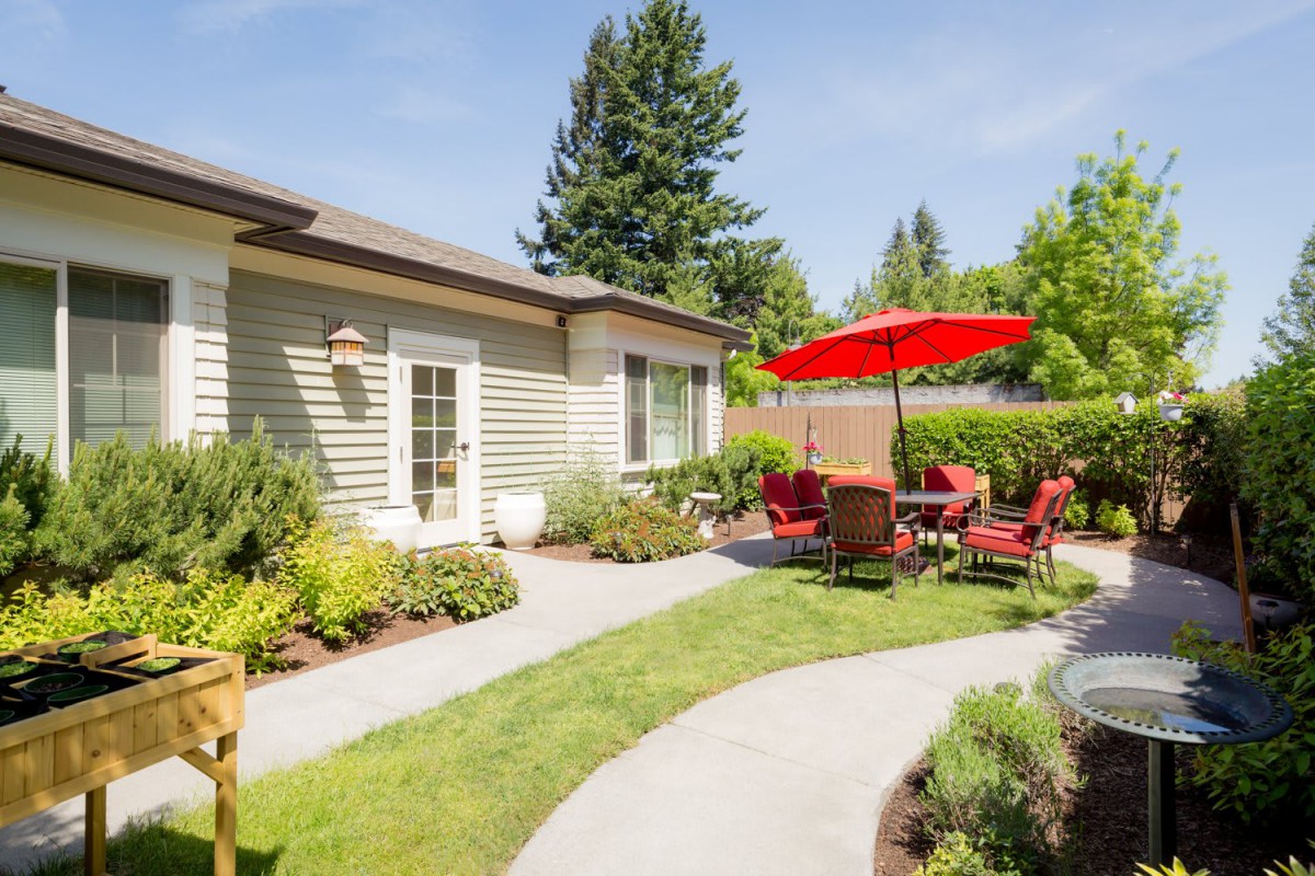 A well-manicured backyard featuring a patio set with a red umbrella, surrounded by lush greenery and a pathway.