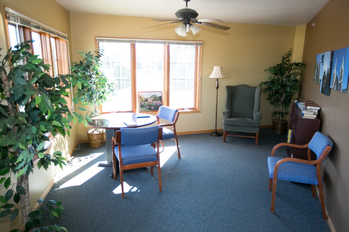 A cozy living room with a table, chairs, armchair, and potted plants near large windows.