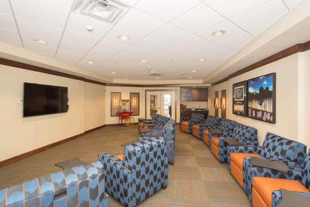 A spacious waiting room featuring patterned chairs arranged along the walls, with framed artwork and a large television mounted on one side.
