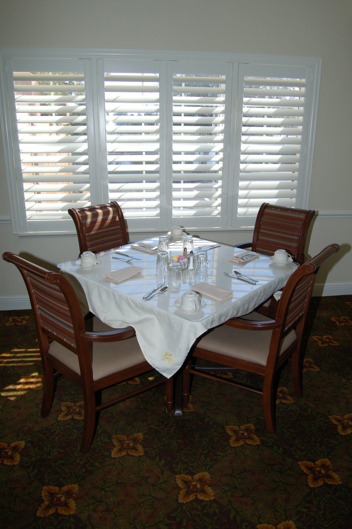 A neatly set dining table for four near a window with shutters.