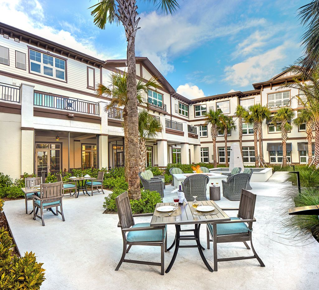 A sunny outdoor patio area with tables and chairs set in front of a modern residential building, surrounded by palm trees.