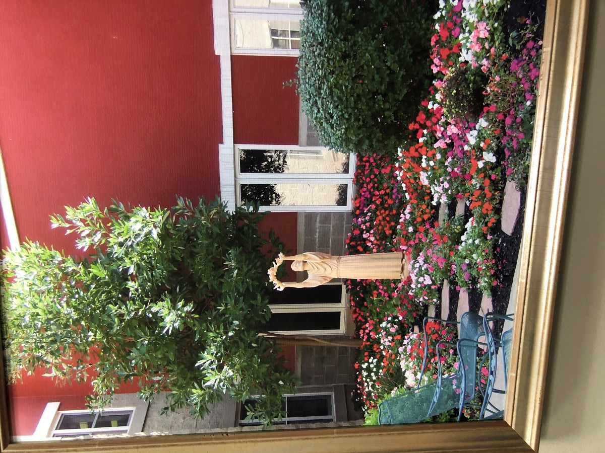 A peaceful courtyard with a statue surrounded by vibrant flowers and greenery against a red wall.