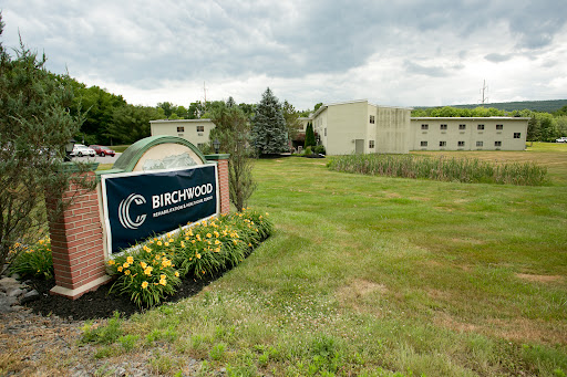 A view of the Birchwood facility surrounded by lush greenery and blooming flowers under a cloudy sky.