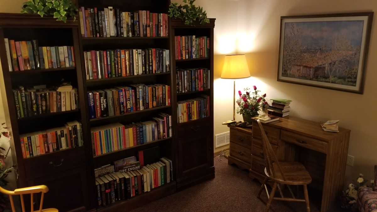A cozy reading corner with a bookshelf filled with books, a desk, a chair, and a warm lamp.