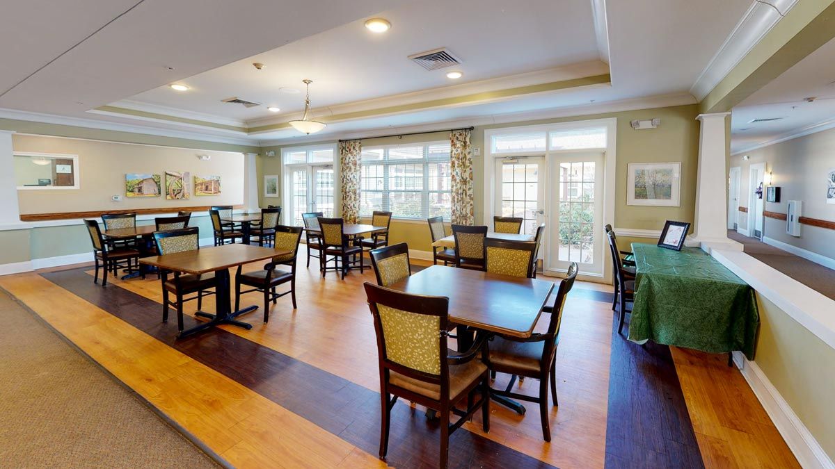 A bright dining area with neatly arranged tables and chairs, featuring large windows and hardwood flooring.