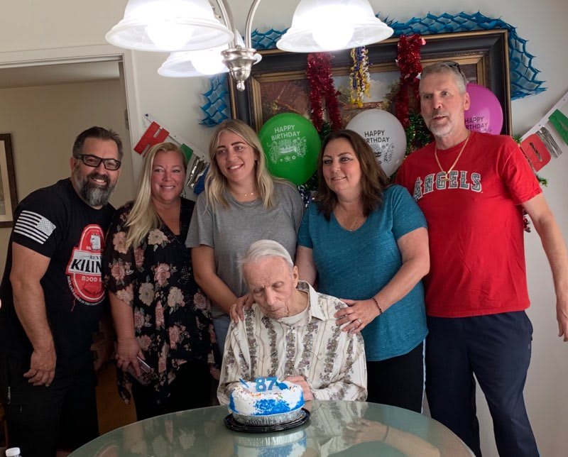 A family gathered around an elderly man celebrating a birthday with cake and balloons.