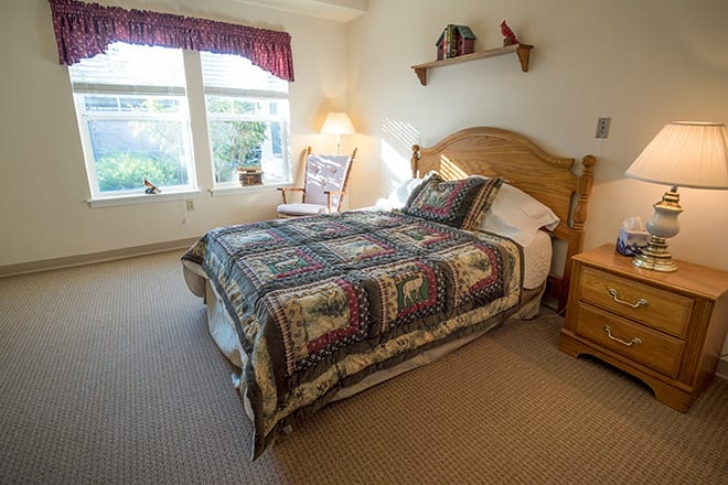 A cozy bedroom with a wooden bed frame and a colorful quilt, featuring natural light from the windows.