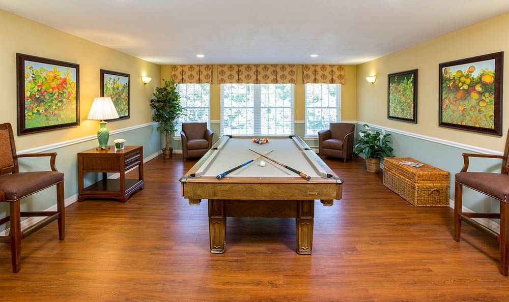 A cozy recreation room featuring a pool table, surrounded by comfortable chairs and large windows, with hardwood flooring and decorative plants.