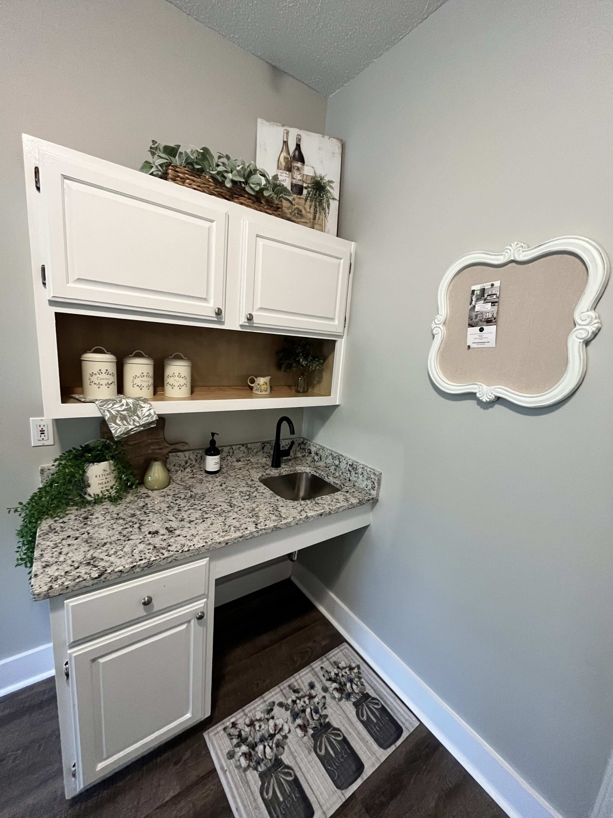 A neatly organized laundry room featuring white cabinets, a granite countertop with a small sink, and decorative greenery.