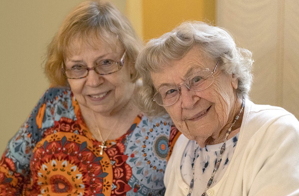 Two elderly women smiling and sitting together indoors.