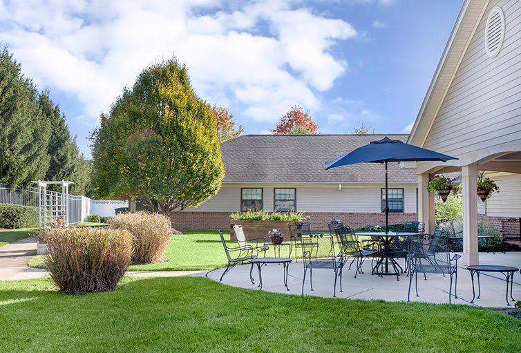 A sunny outdoor patio with furniture and an umbrella, surrounded by a green lawn and trees.
