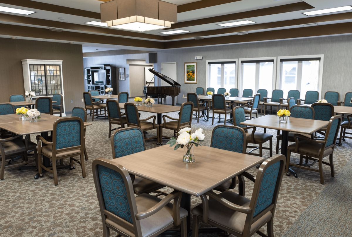 A spacious community dining room with neatly arranged tables and chairs, featuring a grand piano in the corner.