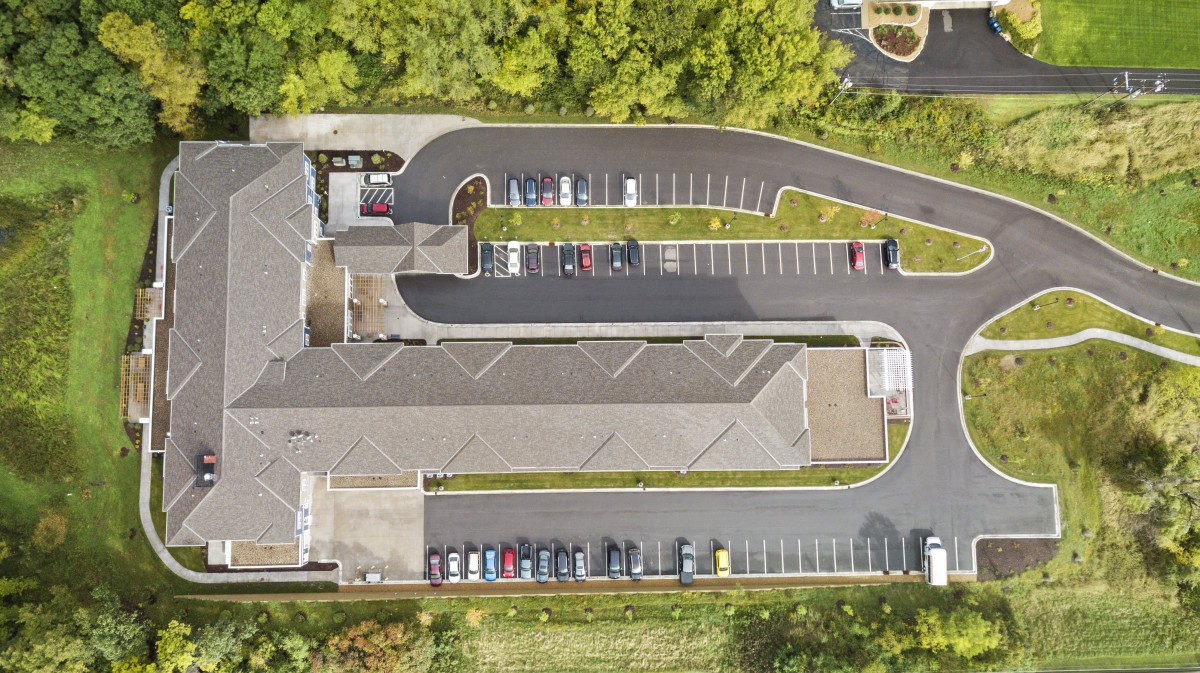 An aerial view of a building with a surrounding parking lot and driveway, bordered by trees.