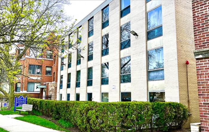 A modern residential apartment building with large windows and a neatly trimmed hedge lining the sidewalk.