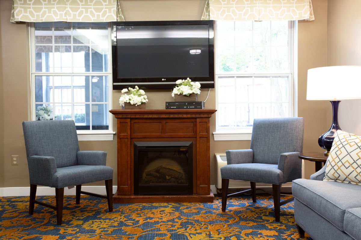 A cozy living room with a fireplace and a TV above, flanked by cushioned chairs and decorated with floral arrangements.
