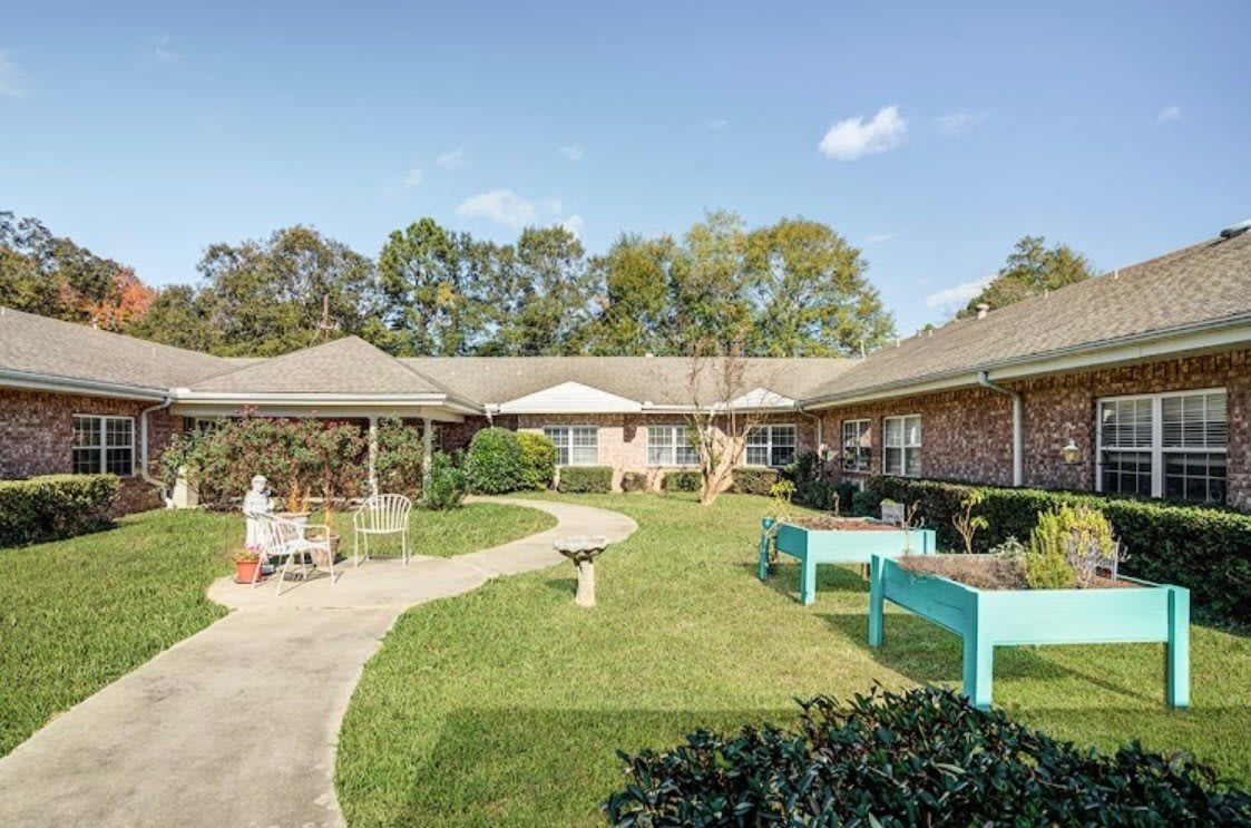 A peaceful courtyard with a winding pathway, surrounded by garden beds and benches, adjacent to a brick building.