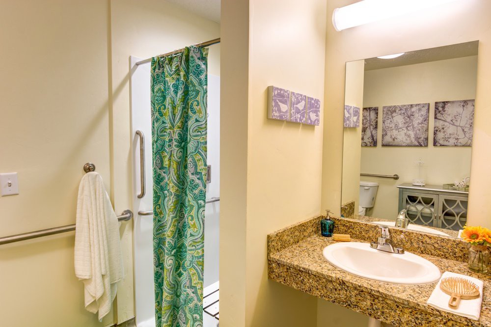 A bathroom featuring a sink with a granite countertop, a shower with a green patterned curtain, and towels hanging nearby.