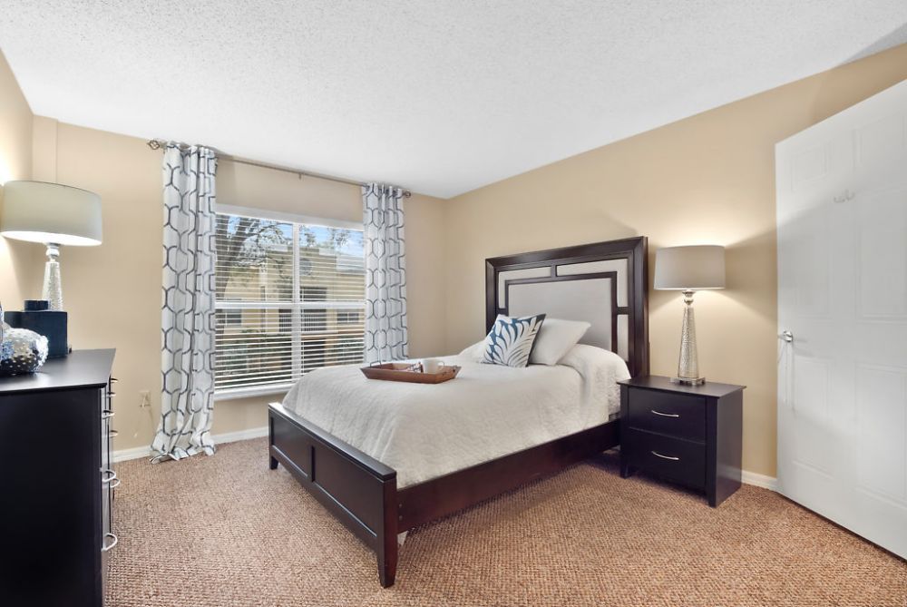 A cozy bedroom with a large window, featuring a bed, lamp, bedside table, and patterned curtains.
