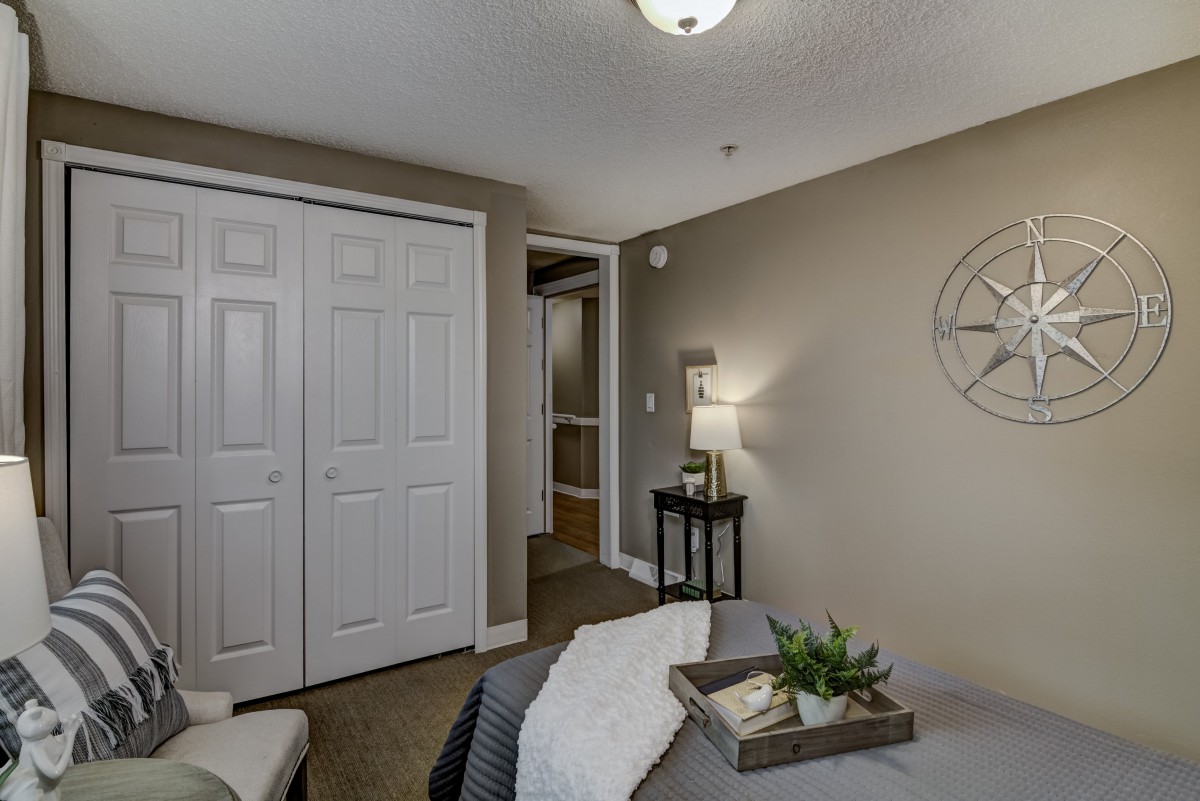 A cozy bedroom with neutral tones featuring a bed, a bedside lamp on a small table, closet doors, and decorative wall art.
