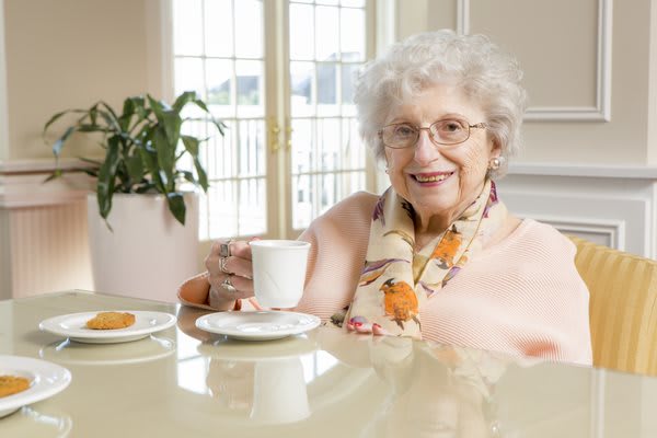 An elderly woman enjoying a cup of tea with cookies at a table.