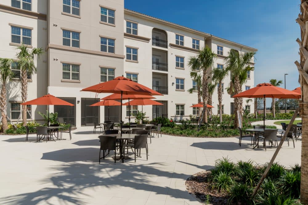 A sunny outdoor patio area with tables, chairs, and orange umbrellas in front of an apartment building surrounded by palm trees.