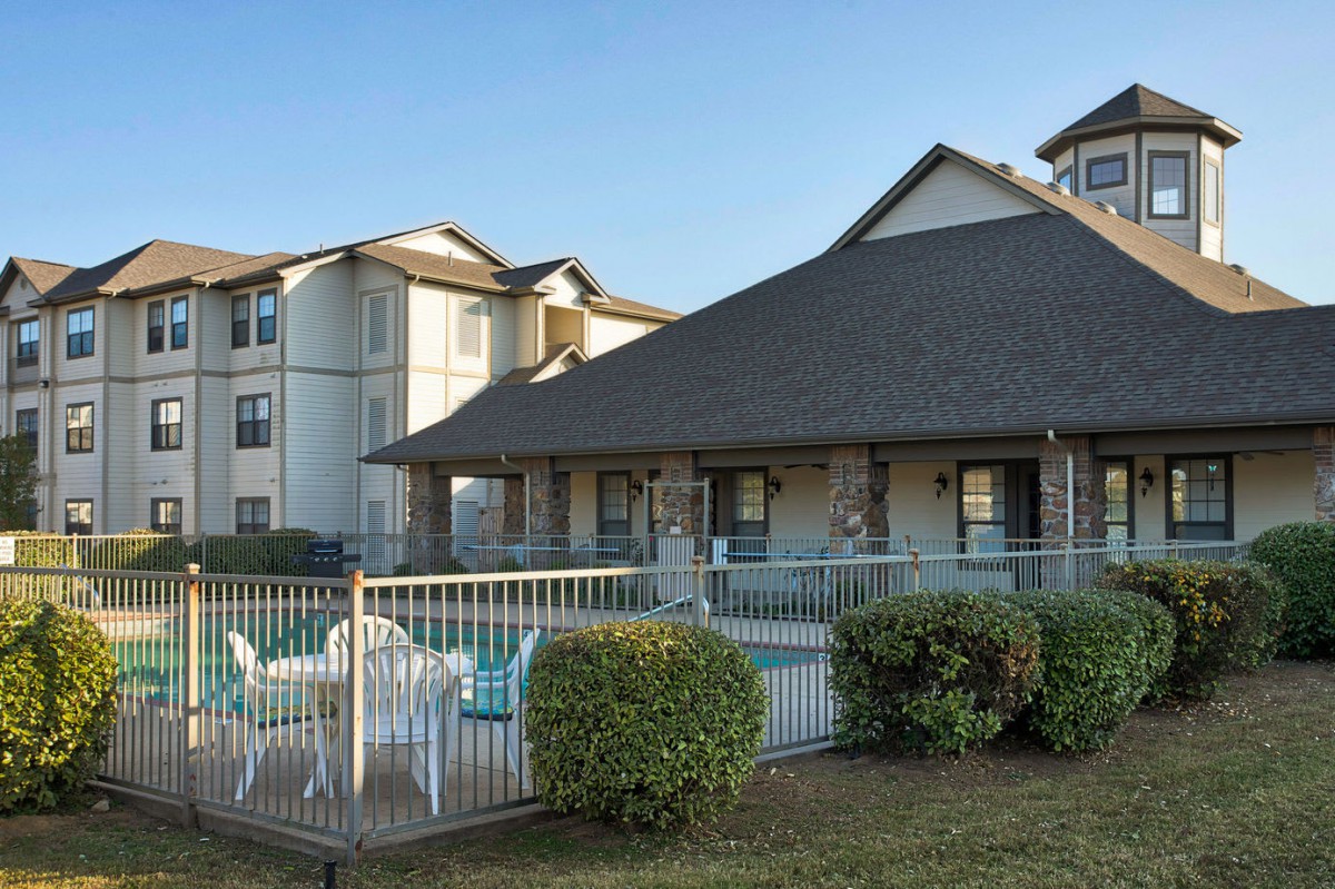 A residential apartment complex with a pool surrounded by a fence and landscaping.