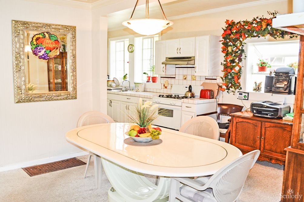 A cozy kitchen and dining area featuring a table with a fruit bowl, surrounded by chairs and decorated with greenery.