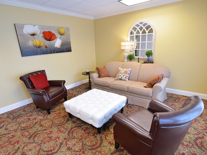A cozy living room with a beige sofa, two leather armchairs, and a white ottoman coffee table. The room is decorated with a colorful artwork on the wall and features warm yellow walls.