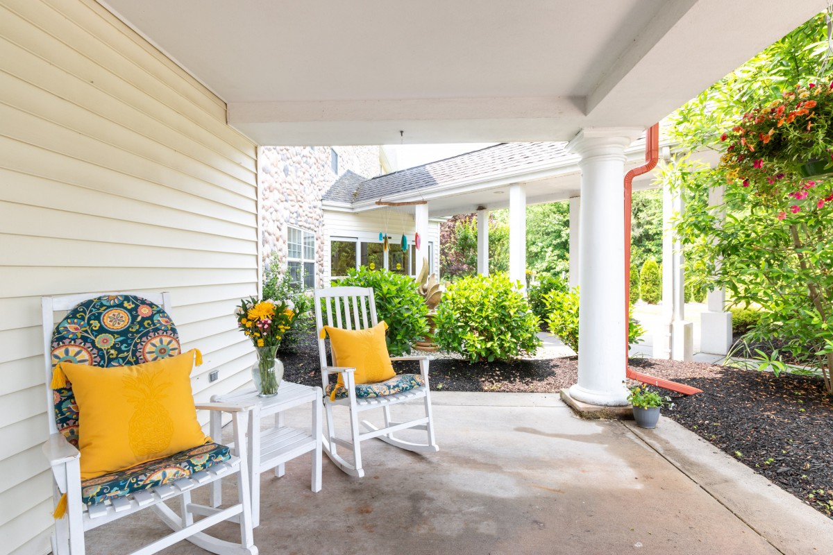 A cozy porch featuring white rocking chairs with yellow pillows, a small table with a vase of flowers, surrounded by lush greenery.