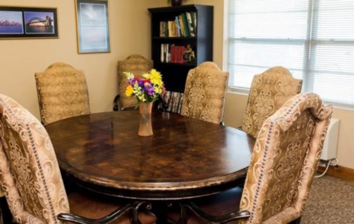 A cozy dining room with a round wooden table and upholstered chairs, featuring a vase of colorful flowers and a bookshelf in the background.