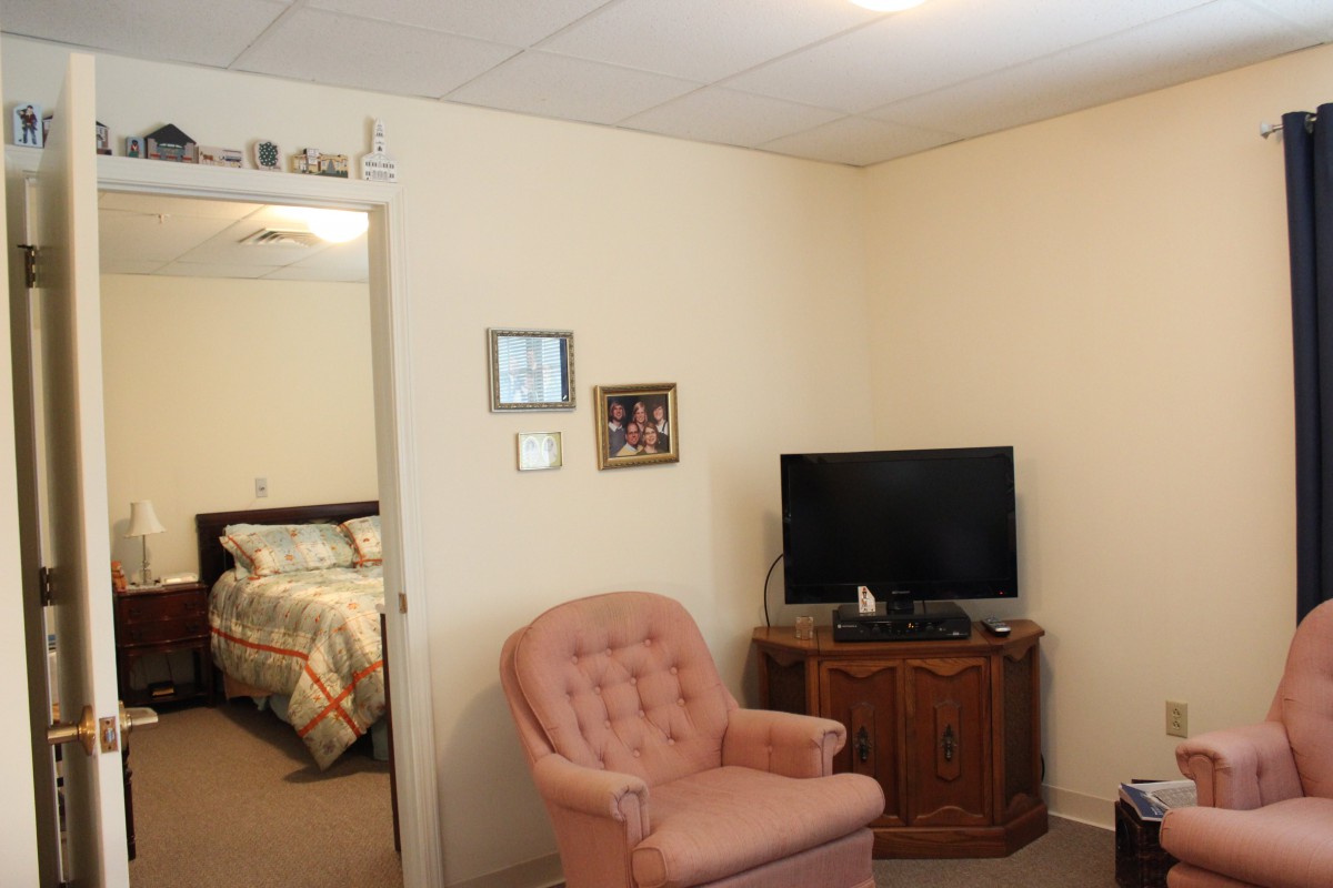 A cozy living room with an armchair and a television, opening up to a bedroom.