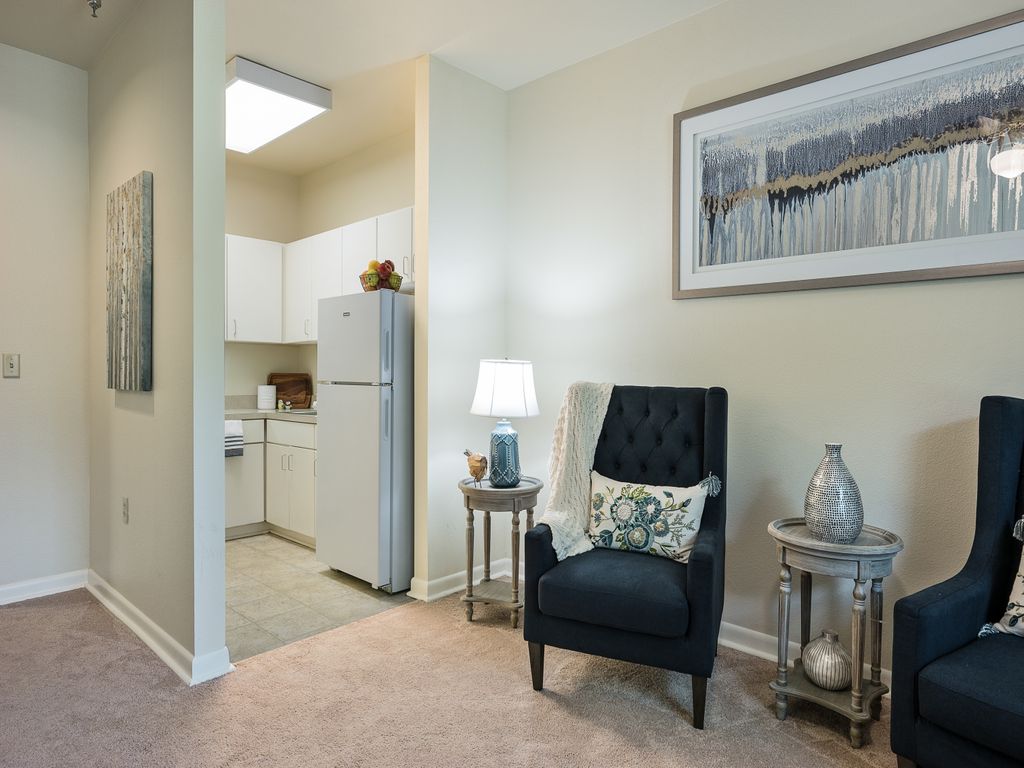 A cozy living area with a view into a small kitchen, featuring a comfortable black chair and side tables with a lamp and decor.