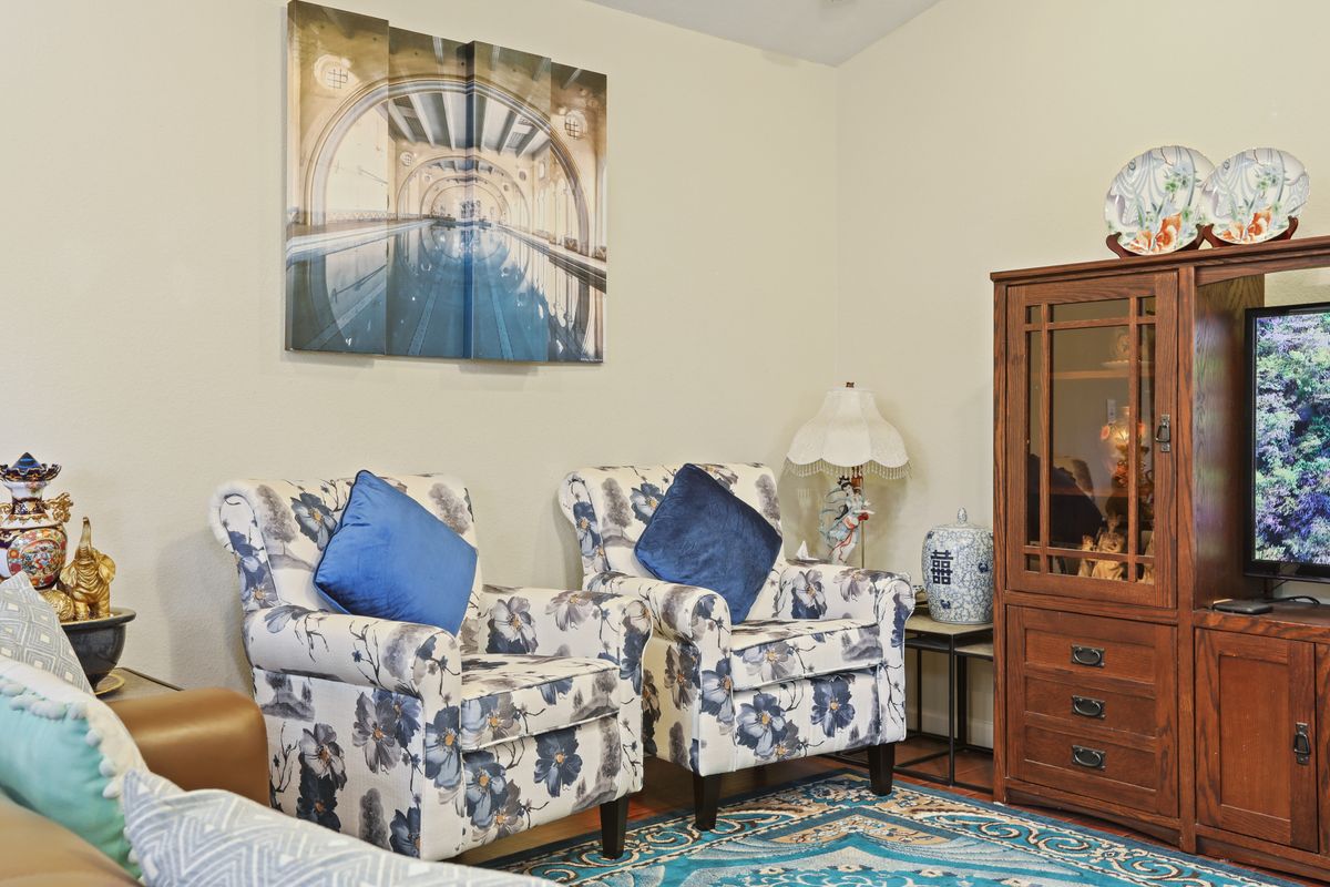 A cozy living room featuring two patterned armchairs with blue pillows, a scenic artwork on the wall, a colorful rug, and a wooden cabinet next to a decorative lamp.