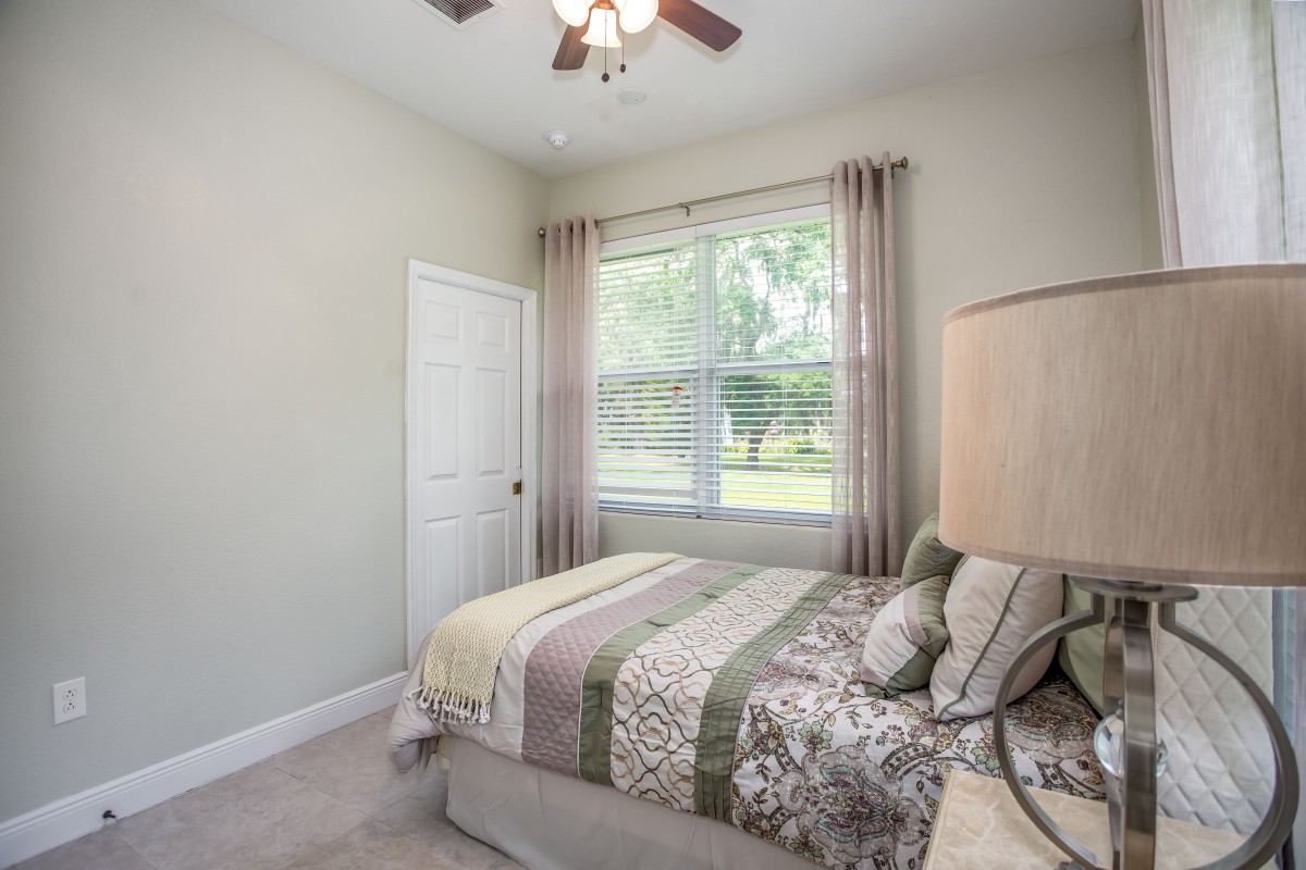 A cozy bedroom with a neatly made bed, a large window with curtains, and a lamp.