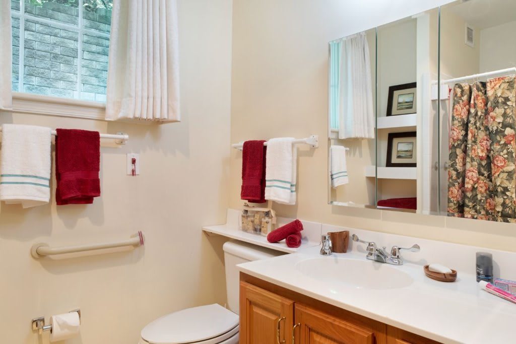 A neatly organized bathroom with a wooden vanity, sink, and large mirror. Red towels add a pop of color to the bright space.