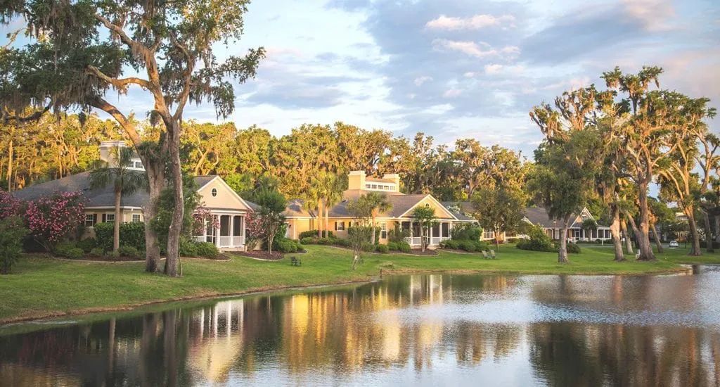 Charming lakeside houses surrounded by trees and reflected in the water under a partly cloudy sky.