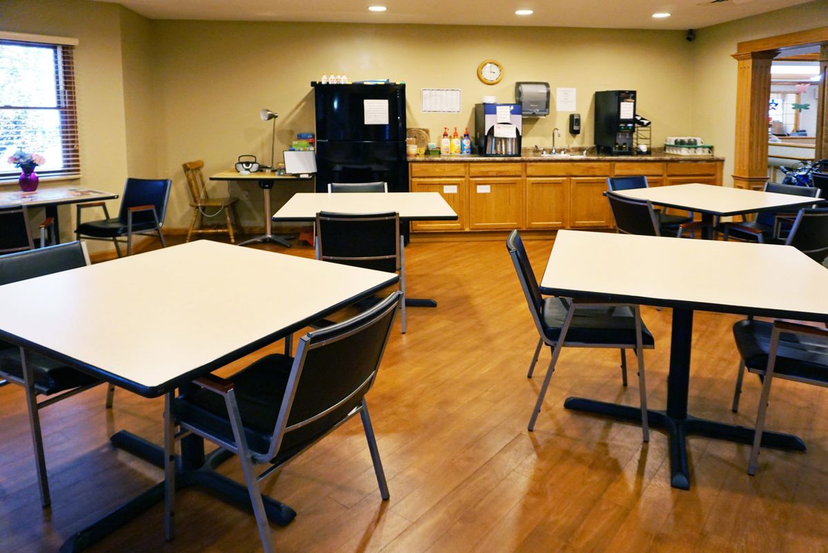 An empty cafeteria with tables and chairs, featuring a small kitchen area in the background.