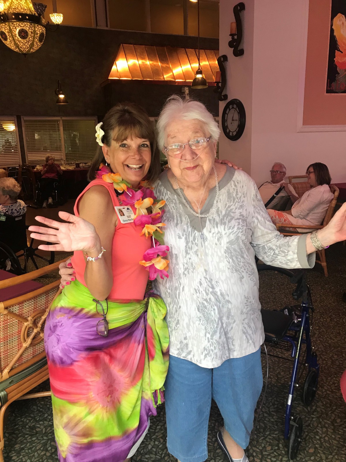 An elderly woman poses with a younger woman dressed in a colorful tropical outfit, both smiling happily indoors.