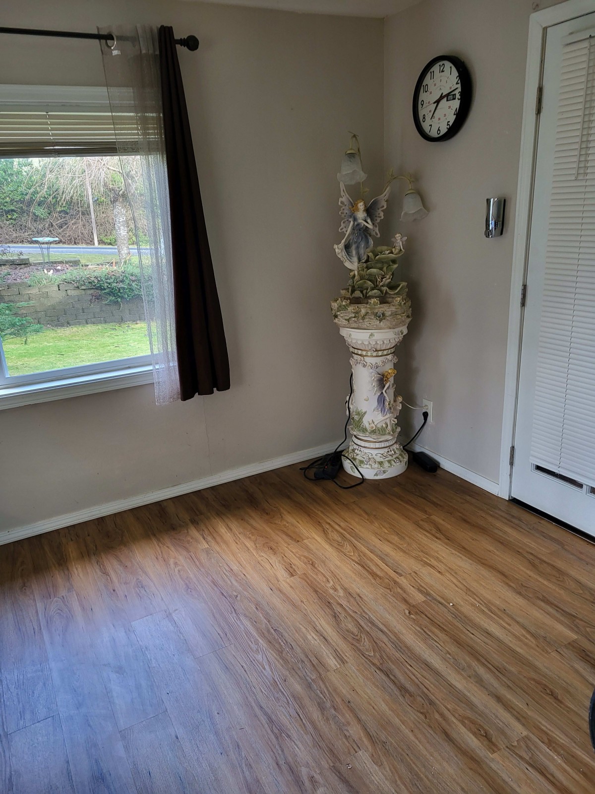 A room with hardwood flooring, featuring a window, a decorative plant stand, and a wall clock.
