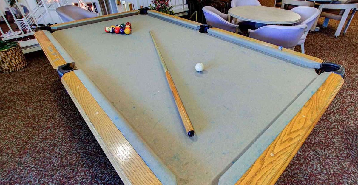 A pool table with a cue stick resting on it and a rack of balls in the background.