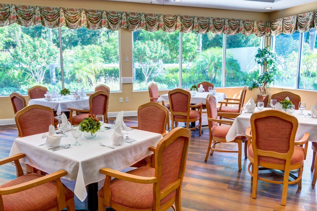 A well-lit dining area with neatly arranged tables and chairs, offering a view of the lush greenery outside through large windows.